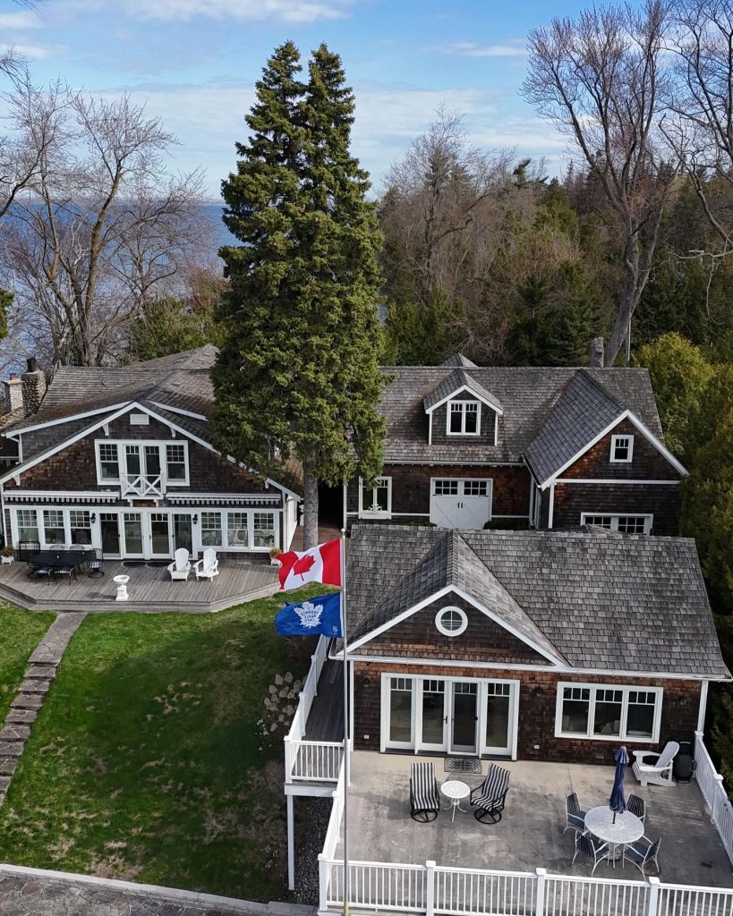 Aerial view of a lakeside house with shingle roofing, a Canadian flag, and a Toronto Maple Leafs flag in a scenic backyard setting.